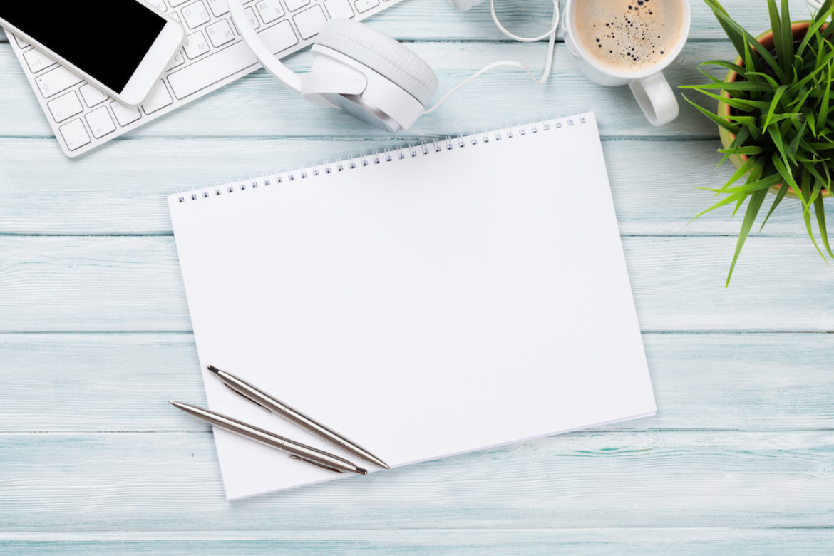 wooden desktop view of notepad, headphones, phone, keyboard, coffee, green plant