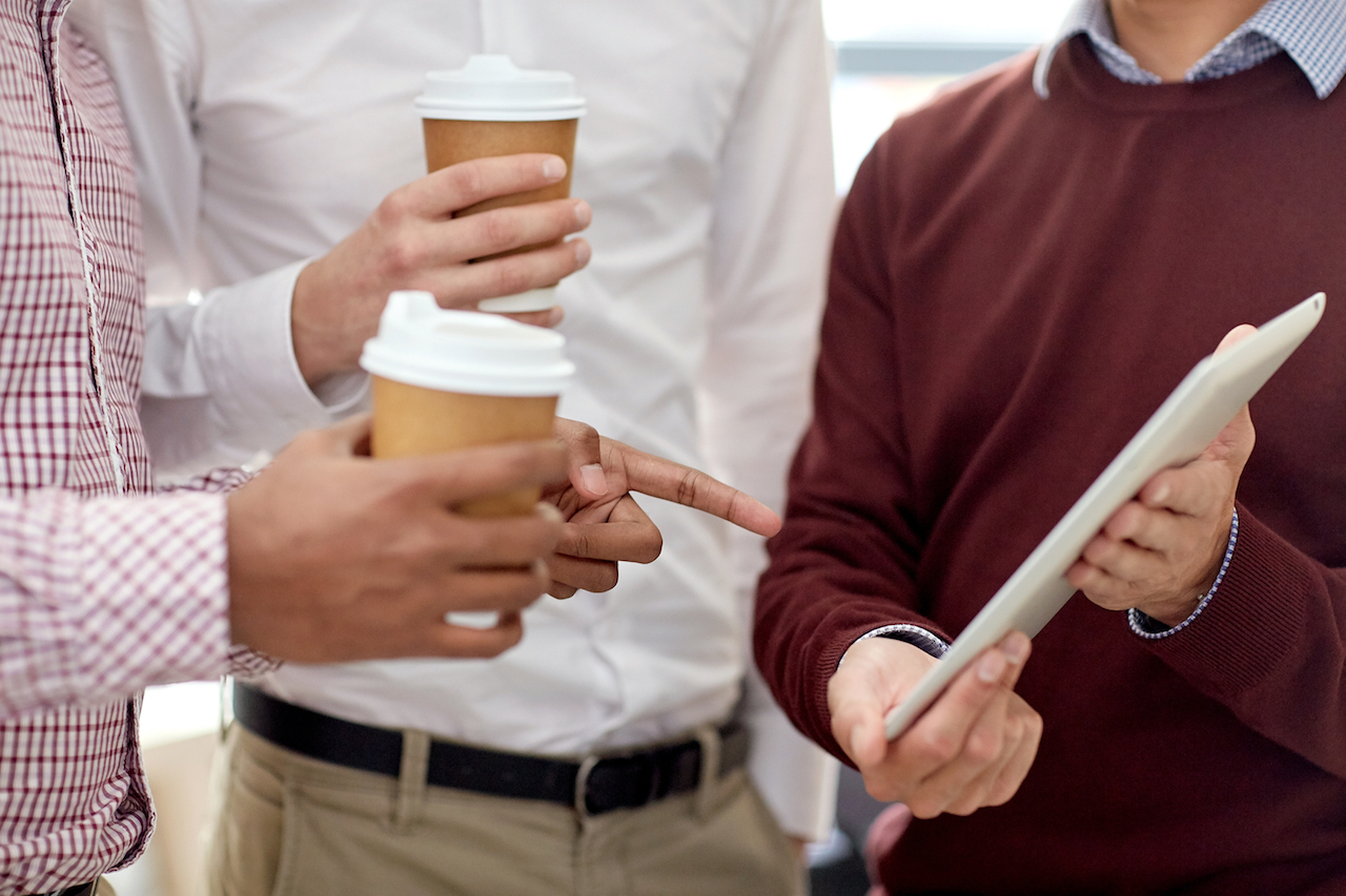 three male team members discuss credits as two hold coffee cups and one holds iPad