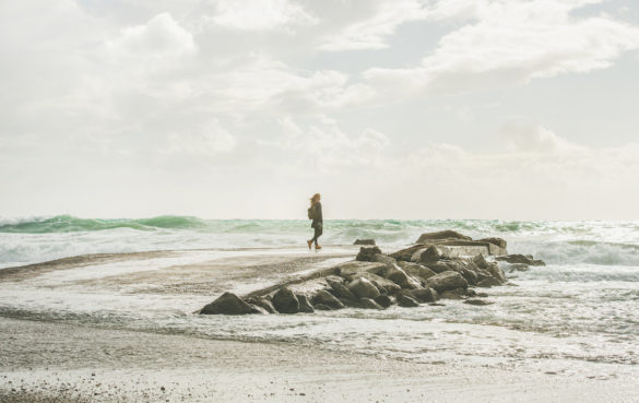 young woman traveler after adversity stands on jetty by rocks near stormy sea