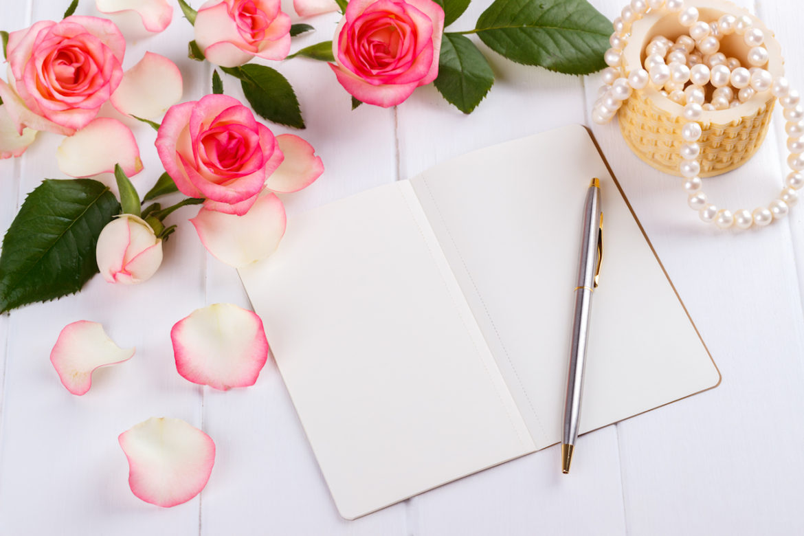 open blank journal with ballpoint pen, pink roses and pearls on white wooden desk, overhead view romantic concept