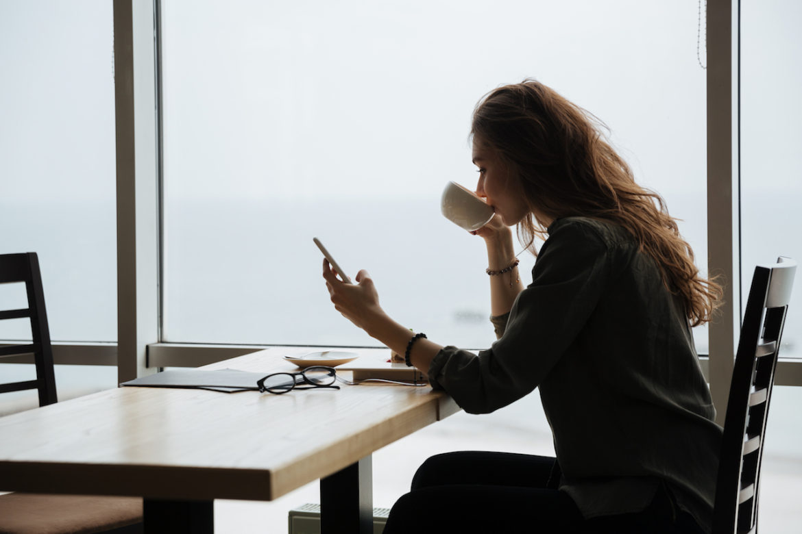 young woman discovering truth while using phone and sipping tea in a cafe by a big window