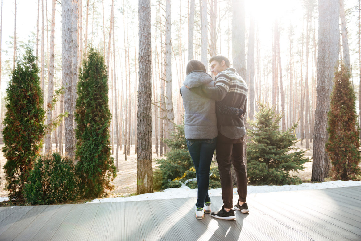 back view of couple who had enough, arms around each other while standing on doorstep facing forest