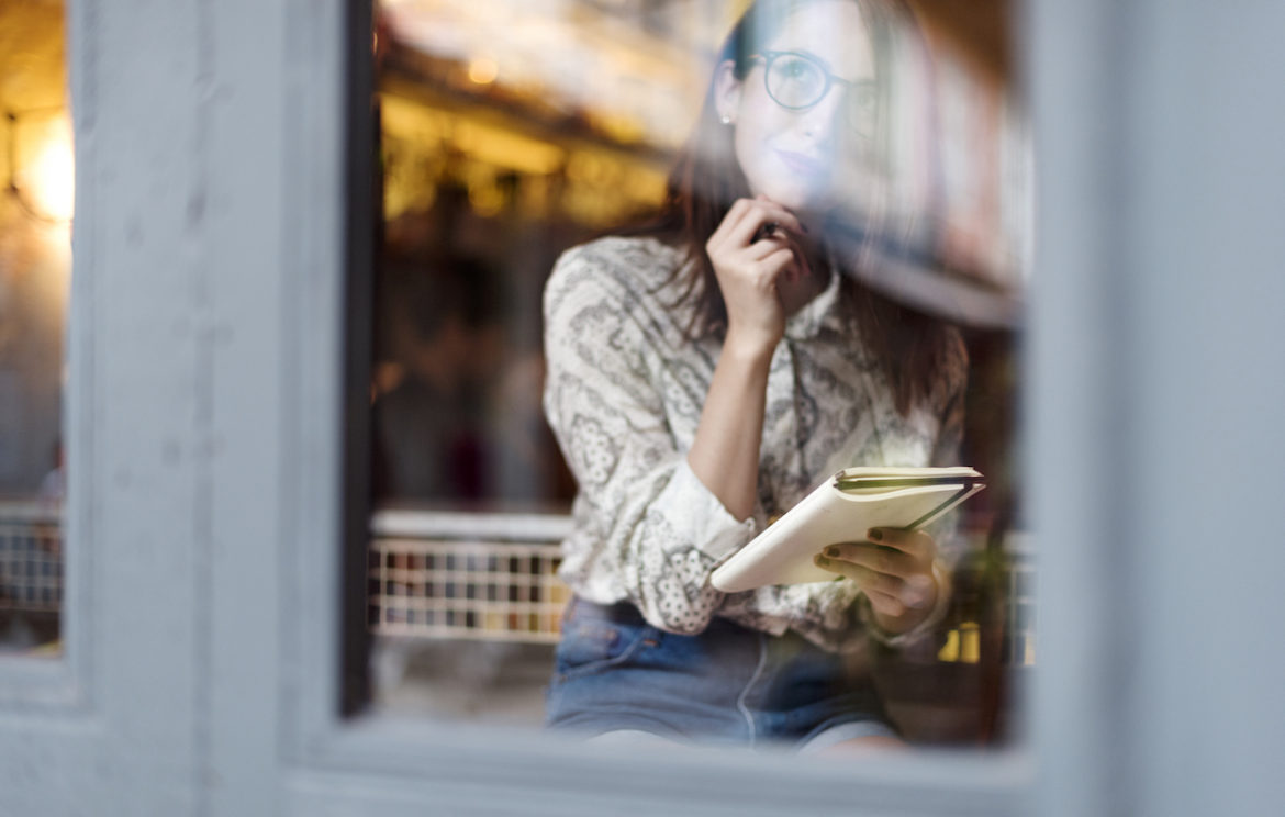 young woman in deep thought with smile creating own rules inside looking out window with notebook in hand