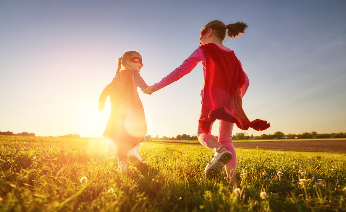 a masquerade played by two young girls holding hands with red masks and capes running on a field at sunset