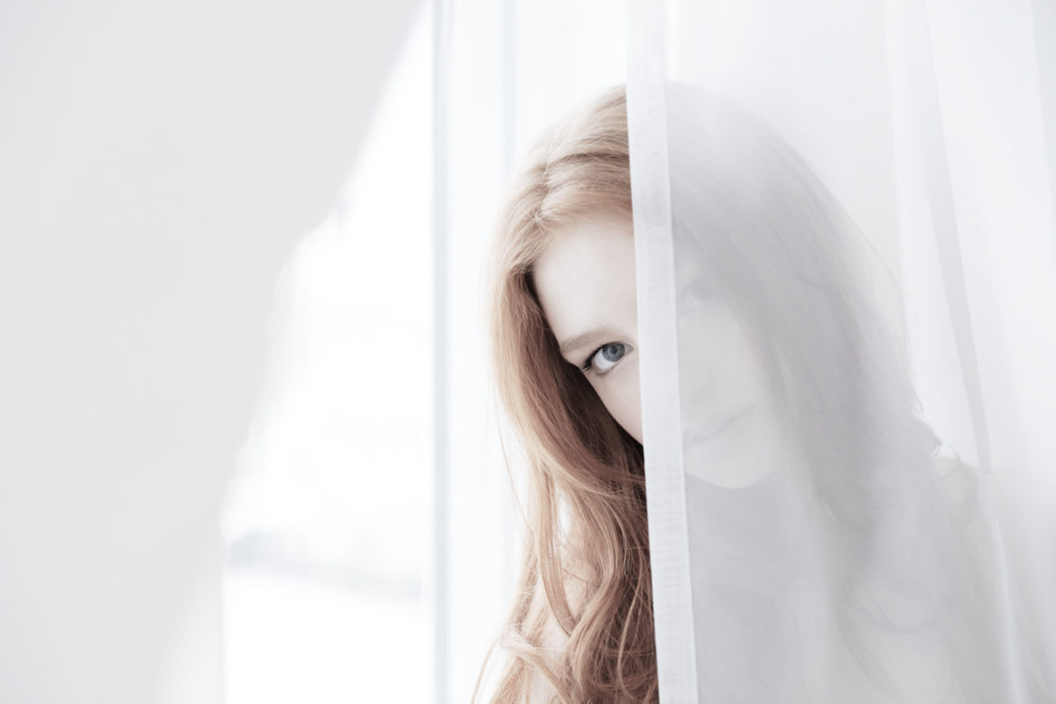 Young woman with long reddish blond hair hides behind white semi-transparent curtain perhaps practicing mindfulness perhaps not.