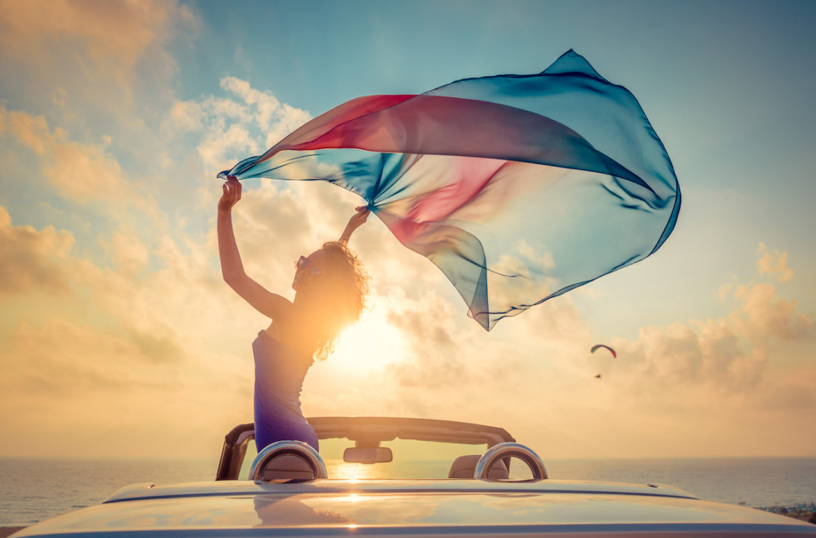 a free spirit in a convertible at the beach waving her multi coloured scarf while friend paragliding from a distance