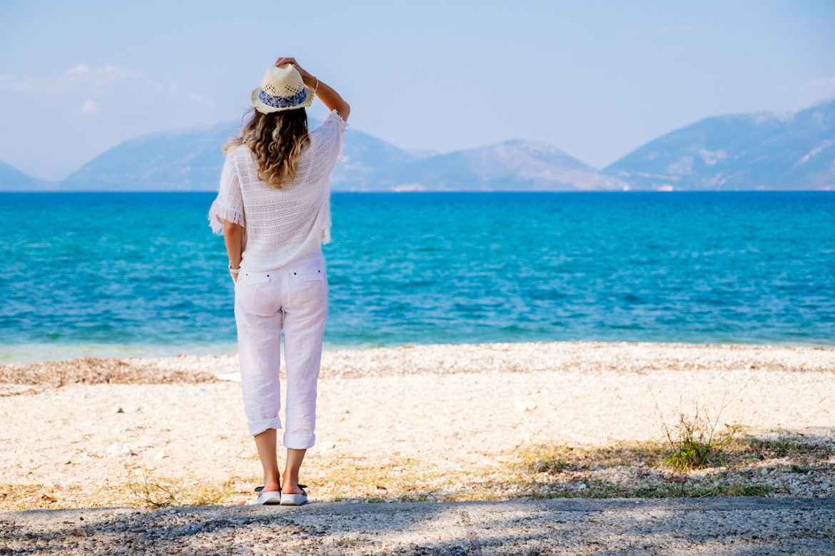woman bringing balance back thinking while holding down straw hat at seaside looking out at the backdrop of hills on a sunny summer day