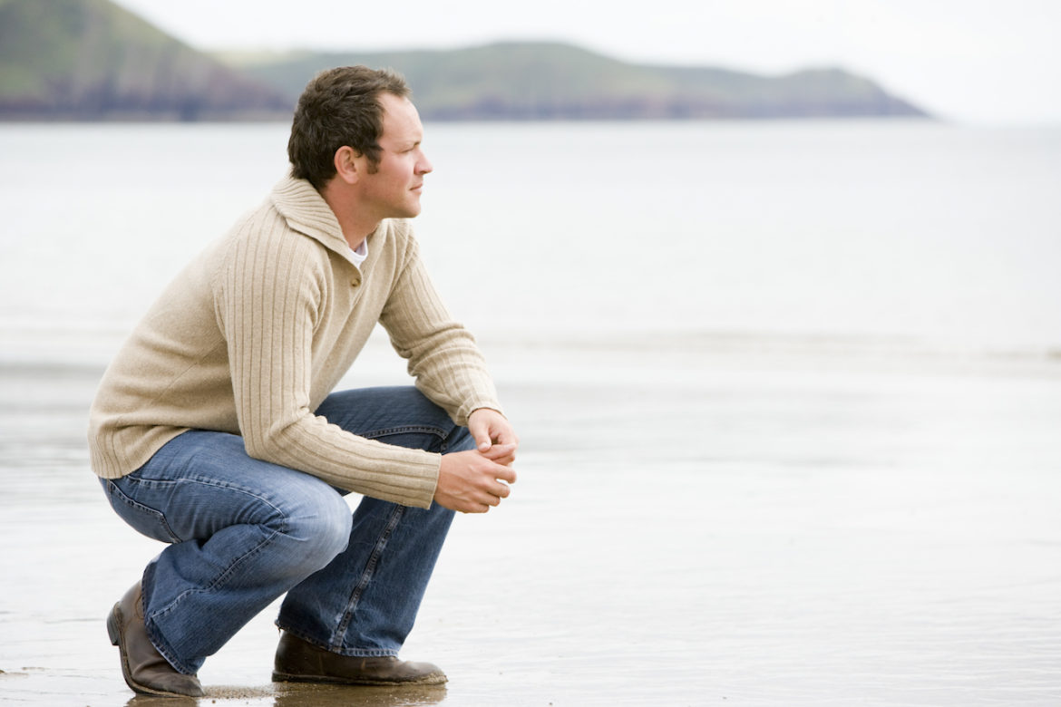 man crouching on the beach accepting closure while looking out beyond the shoreline