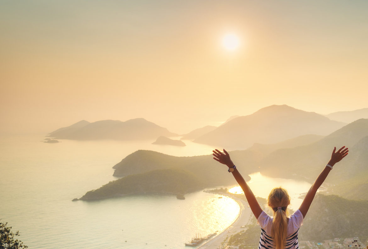From fear to faith woman raises arms standing on a rock looking out at the seashore at sunset in Oludeniz Turkey.
