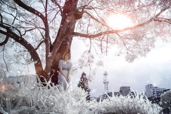 Woman stands with eyes closed exercising the power of intention leaning up against a tree with the sun shining through its branches