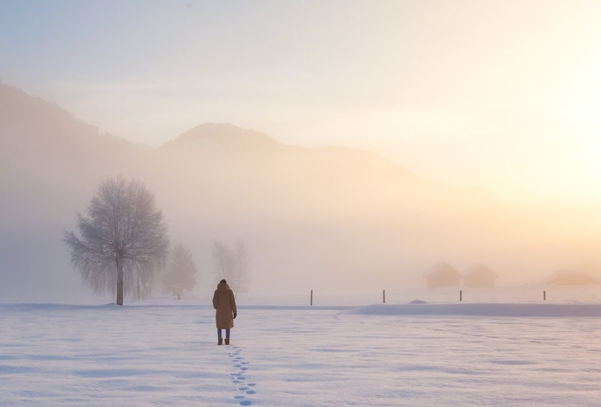 a foggy winter day a woman walks beyond fear toward home leaving footprints trailing behind her in the snow with the sun setting in the horizon