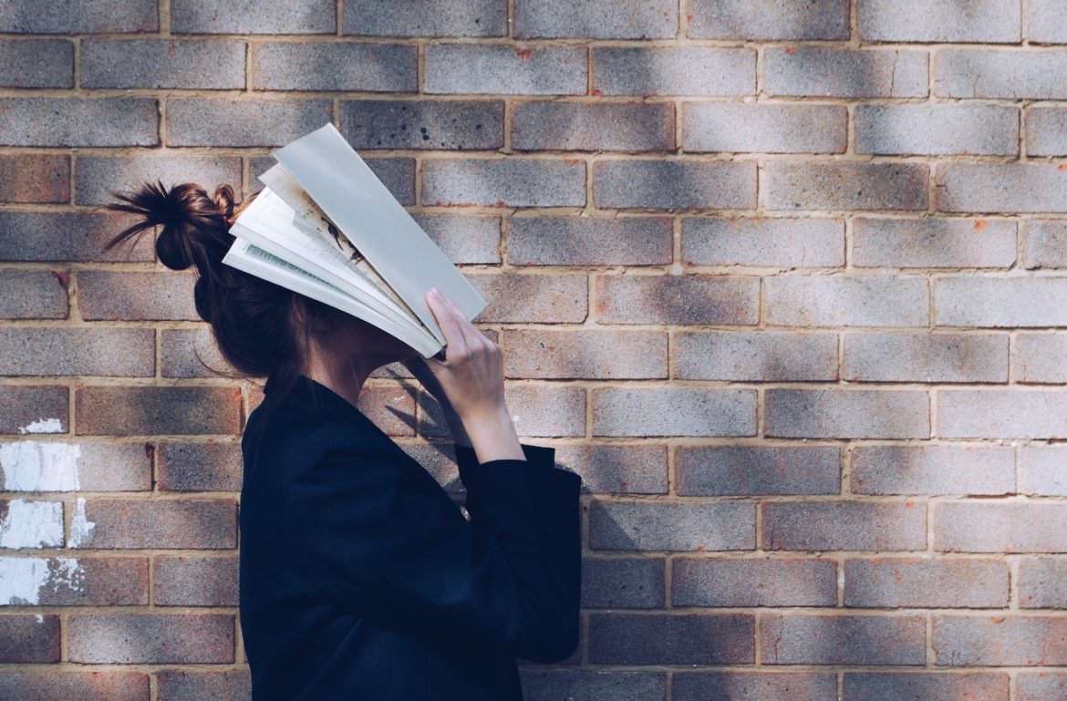 side pose of a woman standing by brick wall facing up with a book closely held to her face reviewing lessons of truth to value self-love