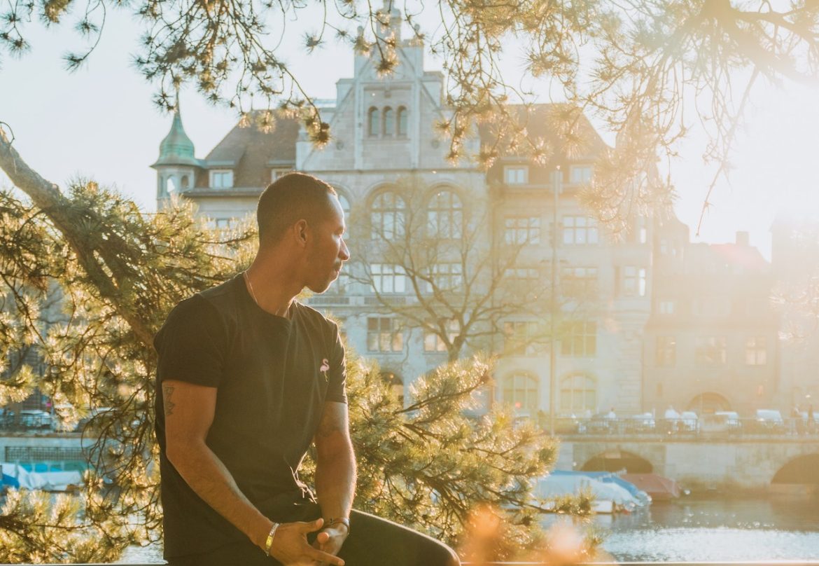 On a sunny day he sits by the canal near a historical site, taking a personal time-out