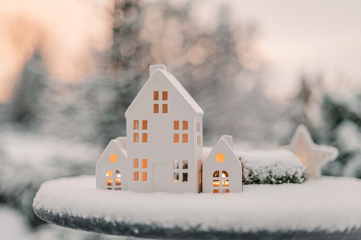 A miniature house with lights perched on a pedestal on a blanket of snow giving one a different perspective at Christmas.